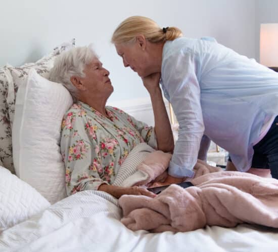 A woman in her 80s in bed with a woman a generation younger bending caringly over her. The implication is that the older woman hasn't been well and the younger woman is taking care of her.