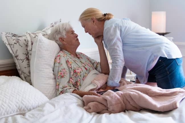 A woman in her 80s in bed with a woman a generation younger bending caringly over her. The implication is that the older woman hasn't been well and the younger woman is taking care of her.