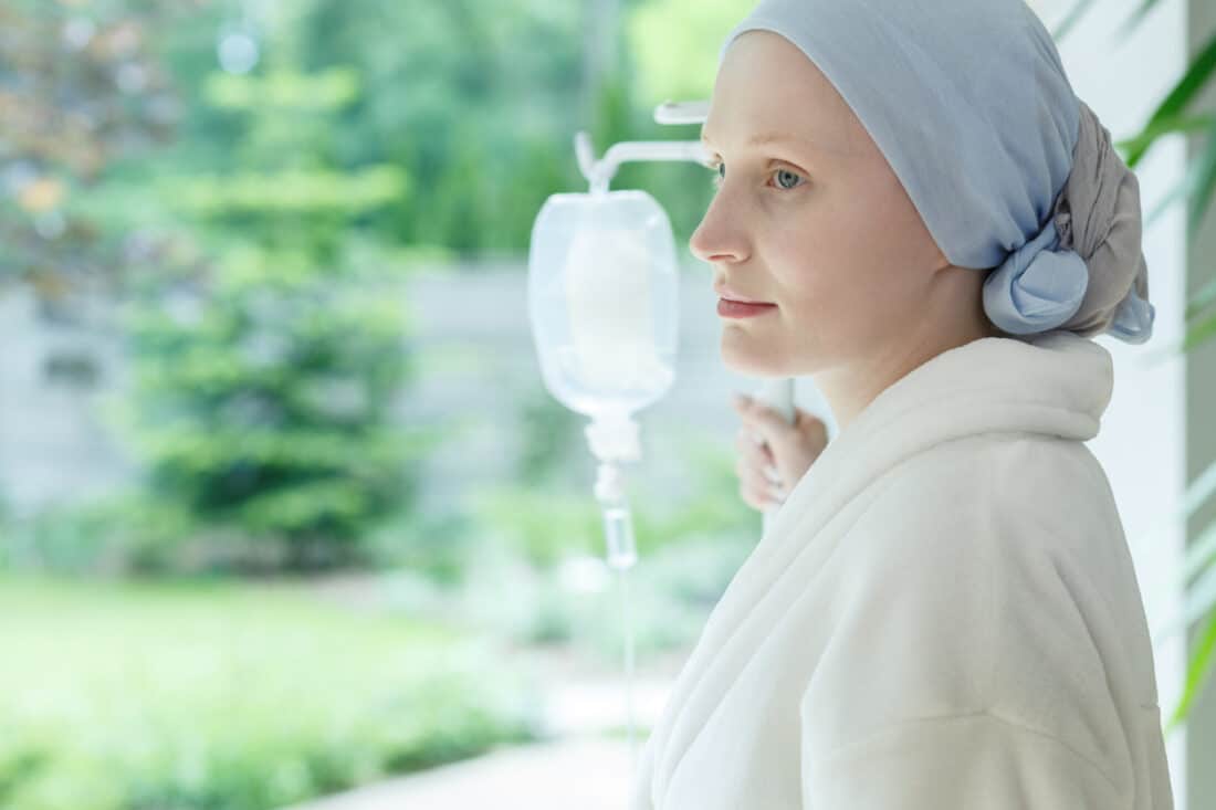 Very pale woman with scarf covering her hair and hospital drip at side, looking out window.