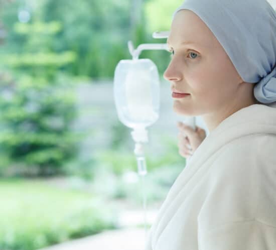 Very pale woman with scarf covering her hair and hospital drip at side, looking out window.